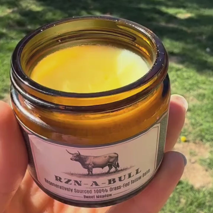 Woman standing in a sunny farm field, smiling as she rubs RZN-A-BULL tallow balm on her hands, praising its silky texture and clean ingredients like jojoba oil, argan oil, and vanilla CO2.