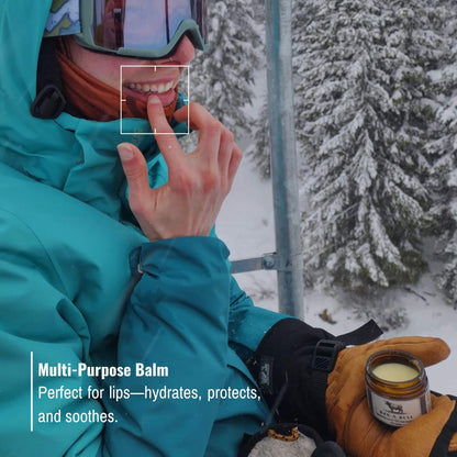 Skier applying Rzn-A-Bull Tallow Balm to lips while on a ski lift in snowy mountains. Multi-purpose balm hydrates, protects, and soothes in harsh winter conditions.
